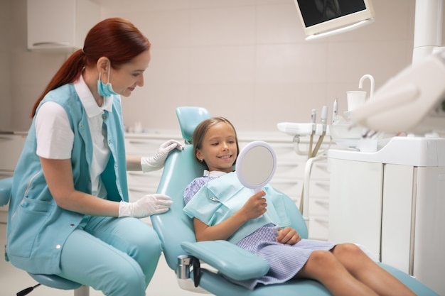 Looking at teeth. Girl looking at her teeth into mirror while visiting dentist in the morning