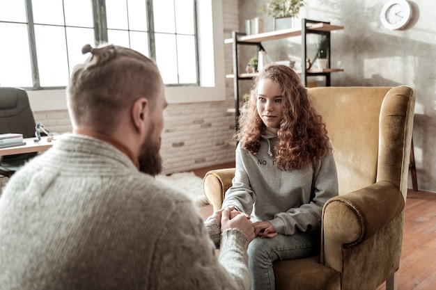 Looking at therapist. Curly appealing teenager wearing grey hoodie looking at her private therapist