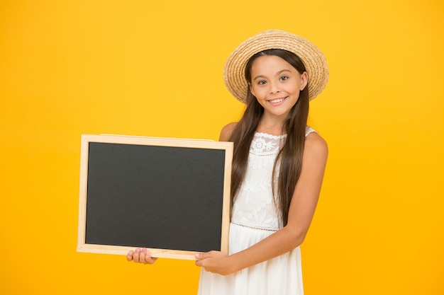 Looking trendy. travel to warm countries. kid spring fashion. girl in straw hat. advertisement. kid traveler . summer shopping sales. happy little girl hold blackboard. copy space. hot vacation time