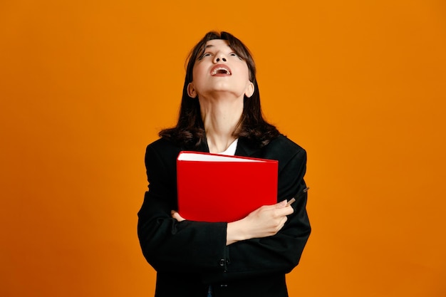 Looking at up holding folder with pen young beautiful female wearing black jacket isolated on orange background