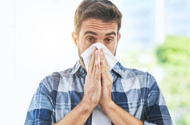looks like its flu season again portrait of an uncomfortable looking young man blowing his nose with a tissue inside of a building during the day