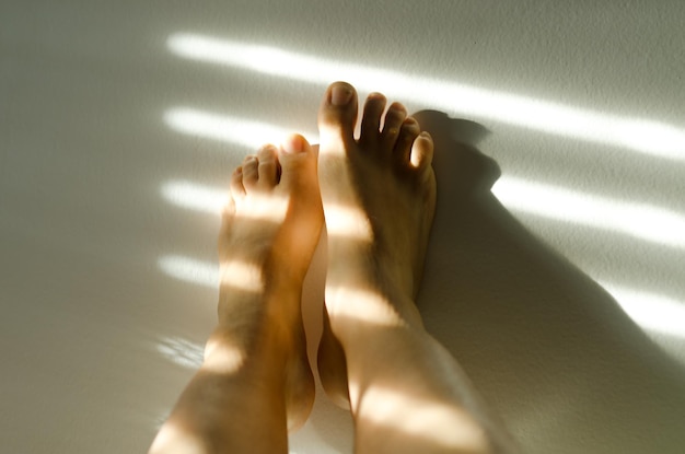 Lose up of a female legs, toes against white wall at cozy home
in sunset light. shadows at the wall.