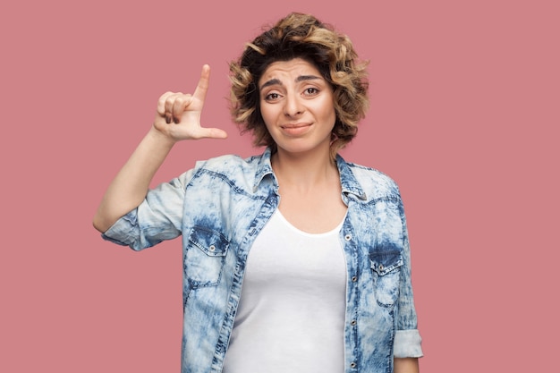 Foto perdente. ritratto di giovane donna triste con l'acconciatura riccia in camicia blu casual in piedi con gesto di perdente e guardando la telecamera con la faccia di fallimento. girato in studio al coperto, isolato su sfondo rosa.