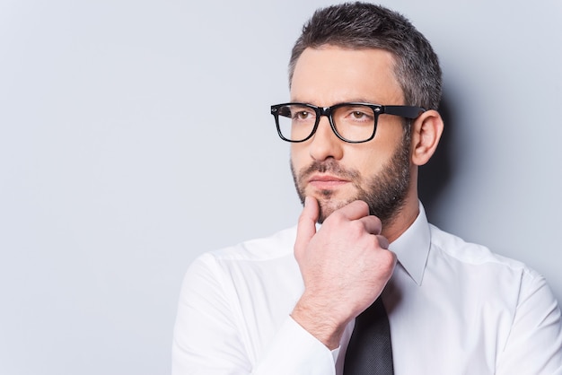 Lost in business thoughts. Portrait of thoughtful mature man in shirt and tie holding hand on chin and looking away while standing against grey background