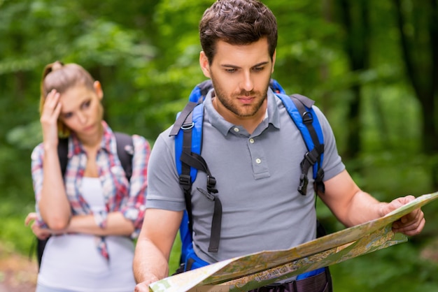 Lost in forest. Thoughtful young man with backpack examining map while woman standing in the background and holding head in hand