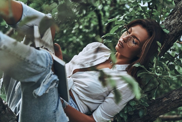 Lost ininteresting book. Attractive young woman in casual wear reading a book while sitting on the tree outdoors