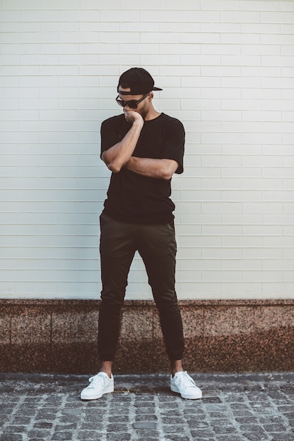 Lost in thoughts. Full length of young mixed race man in casual clothes standing in front of the brick wall and covering face with hand