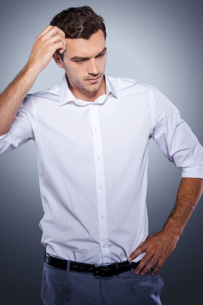 Lost in thoughts. Handsome young man in white shirt holding hand in hair and looking away while standing against grey background