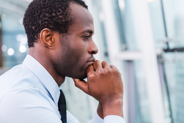 Lost in thoughts. Side view of thoughtful young African man in shirt and tie holding hands on chin