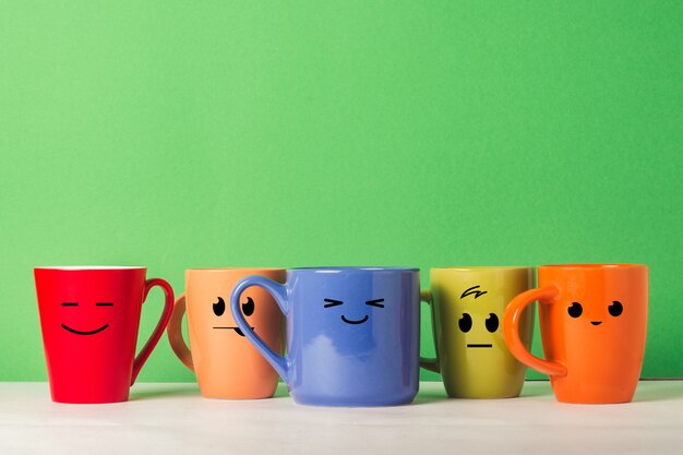 A lot of multicolored cups with funny faces on a green background. The concept of a friendly company, a big family, meeting friends for a cup of tea or coffee, father's day, office, boss day.