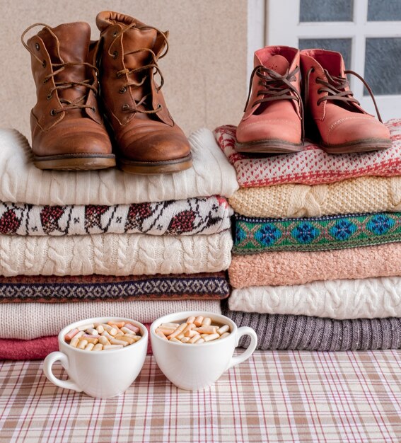 A Lot of Sweaters and Pullovers Different Colours folded in two Piles with brown and red shoes and cups of coffee.