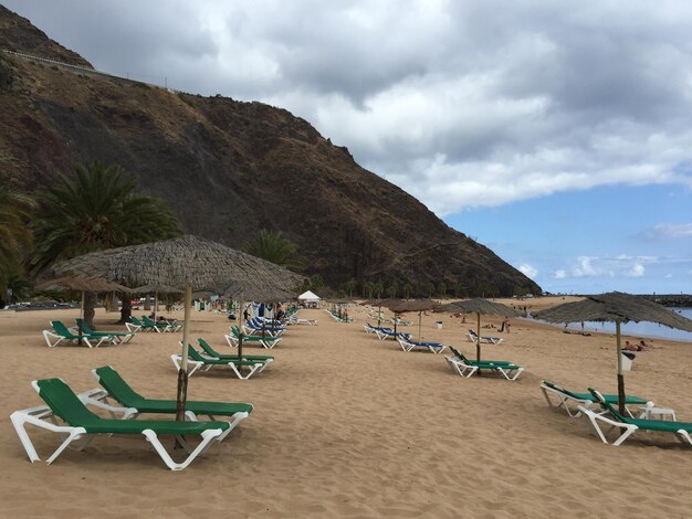 Lounge chairs and parasols on beach by mountain against cloudy sky