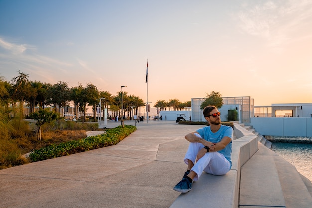 Louvre, Abu Dhabi, United Arab Emirates - May 10, 2020.  Young man sitting by the louvre in Abu Dhabi. Beautiful building at sunset.