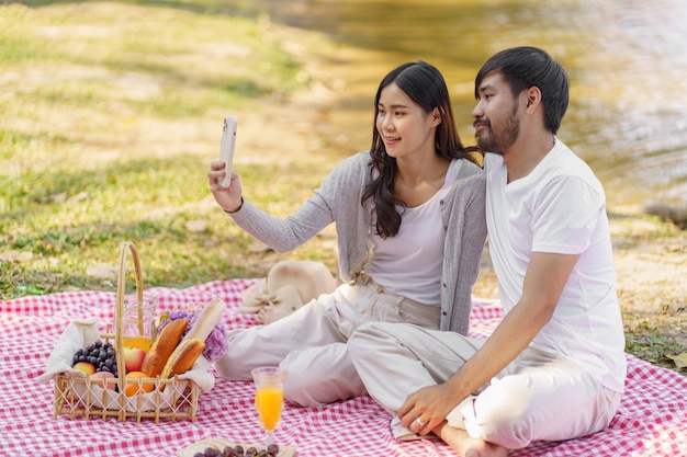 In love asian couple taking selfie photo with smartphone relaxing together with picnic Basket