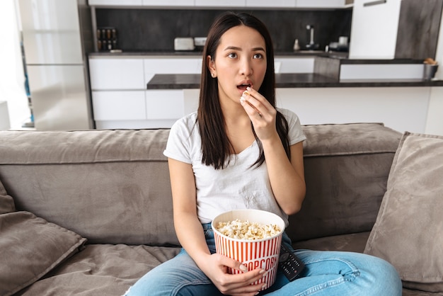 Love asian woman watching TV while sitting on a couch and eating popcorn at home