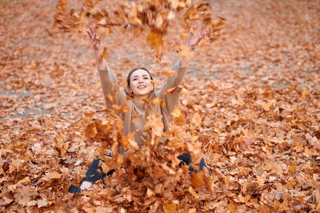 가을을 사랑하십시오. 스웨터, 검은 청바지와 검은 가죽 부츠를 입고 즐거운 매력적인 소녀가 가을 공원에서 단풍을 던지고 있습니다.