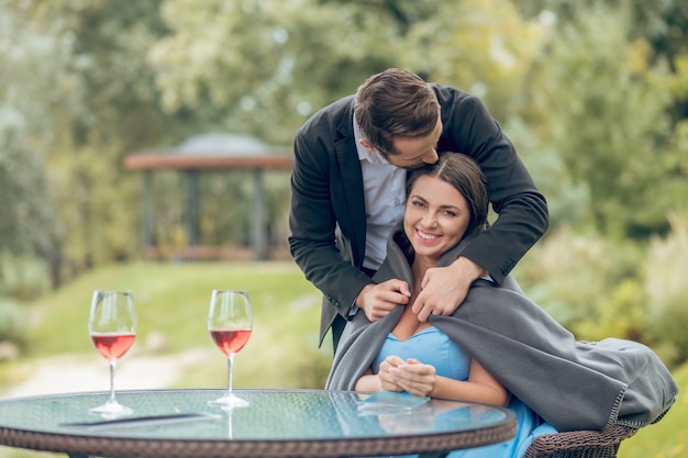 Love, care. Young adult man in dark suit with plaid carefully covering happy pretty woman sitting in cafe on nature