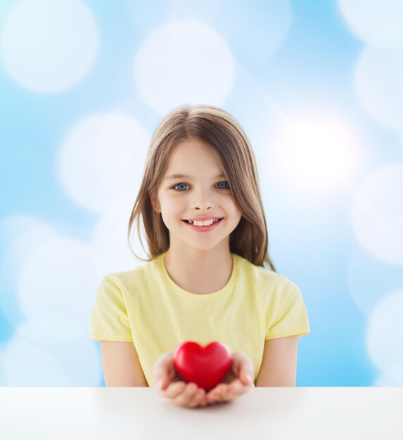 사랑, 자선, 어린 시절, 그리고 사람들의 개념 - 테이블에 앉아 파란 배경 위에 붉은 심장을 들고 있는 아름다운 소녀