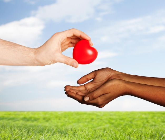 love, charity, valentines day, donation and international concept - close up of man hand giving heart to woman over blue sky and grass background