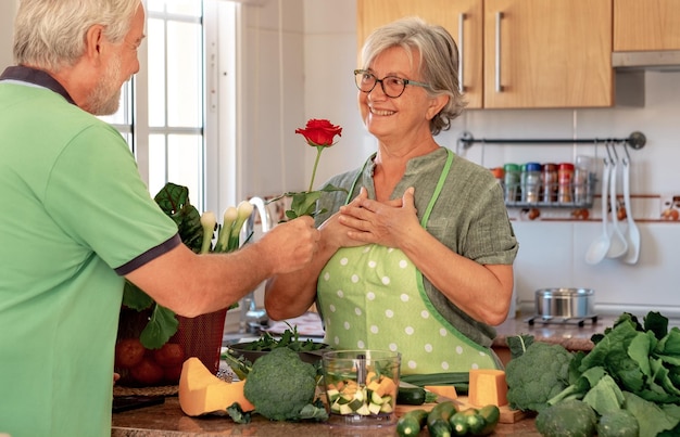 Love concept and valentine\'s day senior bearded husband offers\
his wife a red rose beautiful senior couple in home kitchen while\
preparing vegetables