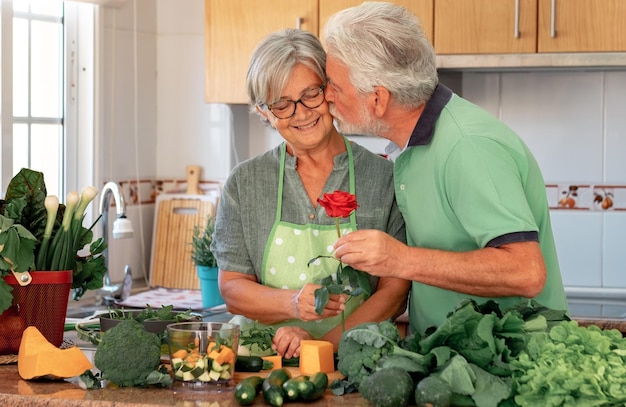 Love concept and valentine\'s day senior bearded husband offers\
his wife a red rose beautiful senior couple in home kitchen while\
preparing vegetables