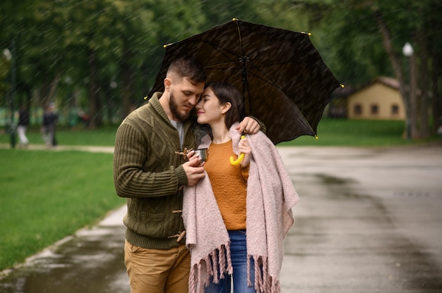 愛のカップルは公園、夏の雨の日にホットコーヒーを飲みます。男と女は雨の傘の下に立つ、散歩道のロマンチックなデート、路地の雨天