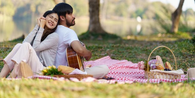 Photo in love couple enjoying picnic time playing guitar in park outdoors picnic happy couple relaxing together with picnic basket