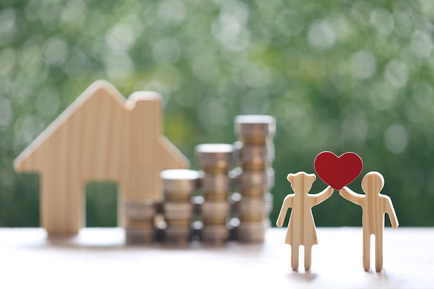 Влюбленная пара держит форму сердца и стопку монет денег с модельным домом на естественном зеленом фоне