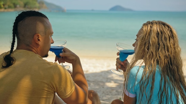 The love couple is holding a glass of blue curacao cocktail on\
sea man with plait and blonde woman drink alcohol on sand beach in\
shadow by azure ocean of a tropical island summer holiday\
concept