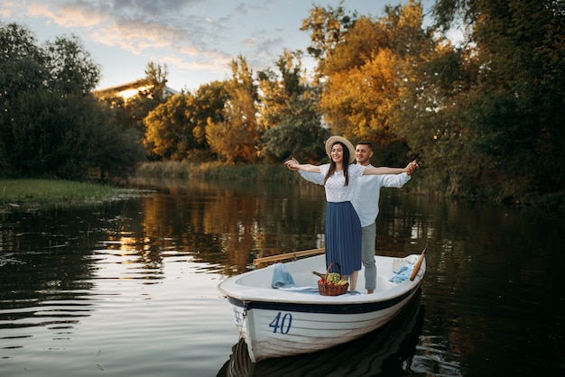 Love couple standing in a boat on quiet lake at summer day on sunset. Romantic meeting, boating trip, man and woman walking along the river