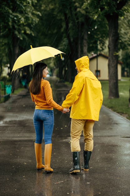 Ami le coppie con l'ombrello che cammina nel parco estivo in una giornata piovosa. uomo e donna in stivali di gomma sul percorso a piedi, tempo umido in vicolo