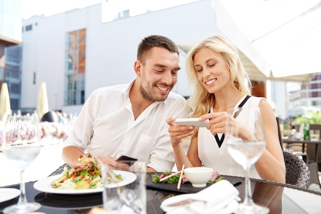 愛、日付、技術、人と関係のコンセプト - レストランのテラスで smatphone と幸せなカップルの笑顔