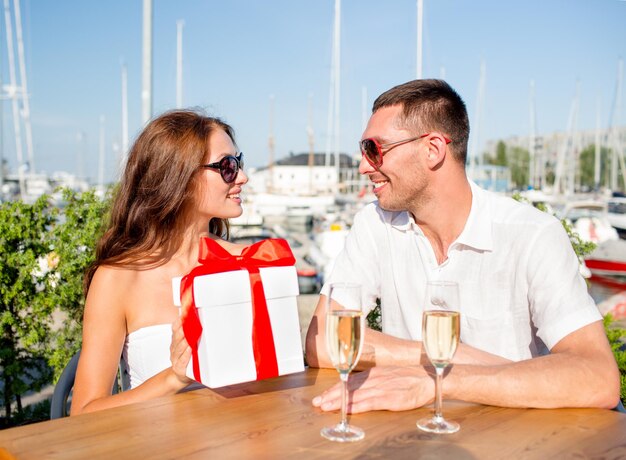 Love, dating, people and holidays concept - smiling couple wearing sunglasses sitting with gift box, flowers and champagne at cafe