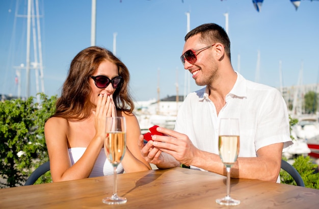 love, dating, people and holidays concept - smiling couple wearing sunglasses with champagne and small red gift box looking to each other at cafe