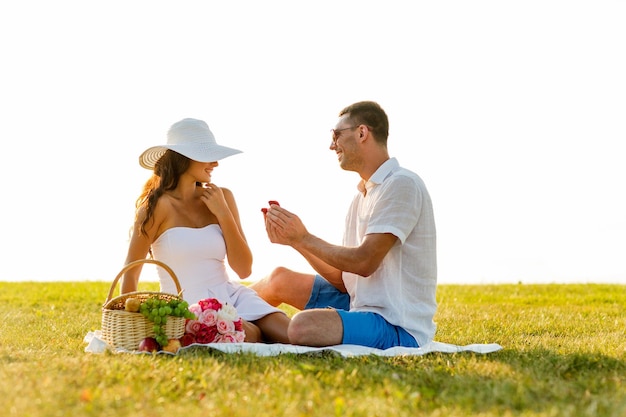 Concetto di amore, appuntamenti, persone e vacanze - giovane sorridente che mostra una piccola confezione regalo rossa alla sua ragazza durante il picnic