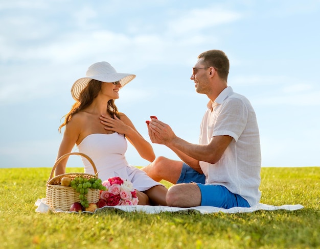 Love, dating, people, proposal and holidays concept - smiling\
young man giving small red gift box with wedding ring to his\
girlfriend on picnic over blue sky and grass background