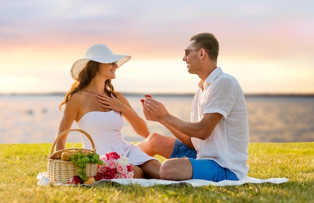 love, dating, people, proposal and holidays concept - smiling young man giving small red gift box with wedding ring to his girlfriend on picnic over seaside sunset background