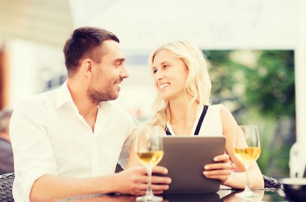 love, dating, people, technology and holidays concept - happy couple with tablet pc computer and glasses of wine at restaurant lounge or terrace