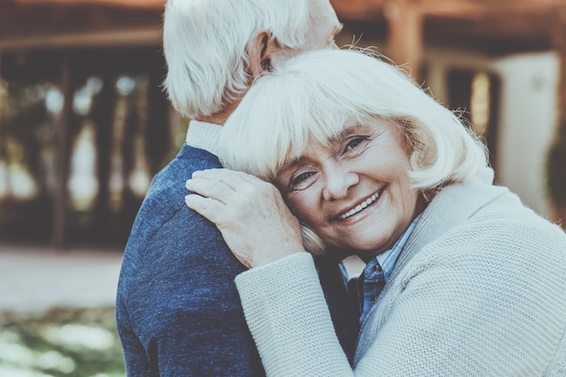 Love forever. Happy senior woman leaning at shoulder of her husband and smiling while both standing outdoors and in front of their house