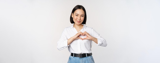 Love gesture Beautiful asian young woman showing heart gesture and smiling express care and affection standing over white background