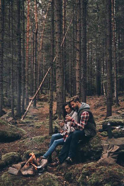 Love is in the air. Beautiful young couple holding cups while warming up near the campfire