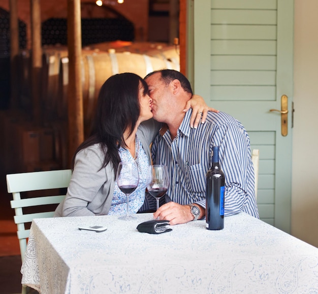 Love kiss and a couple on a wine farm together in celebration of love or their anniversary Kissing romance or dating with a loving man and woman at a table in a winery feeling happy while bonding