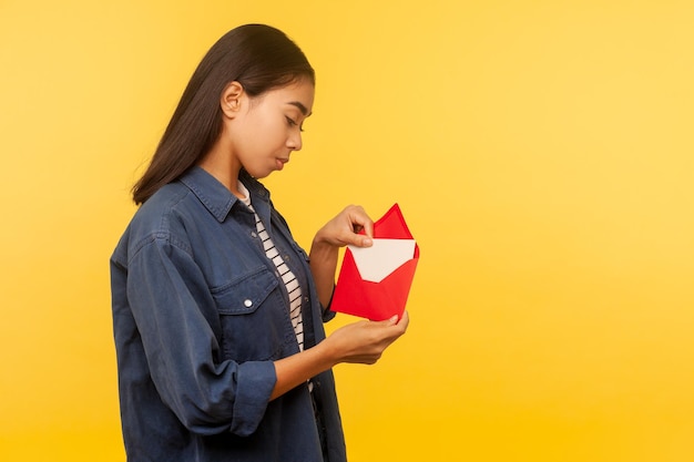 Love letter on Valentine39s day Portrait of girl in denim shirt pulling letter from red envelope holding greeting card and smiling with dreamy expression studio shot isolated on yellow background