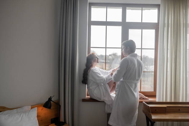 In love. A man and a woman in white bathrobes standing near the window and feeling in love