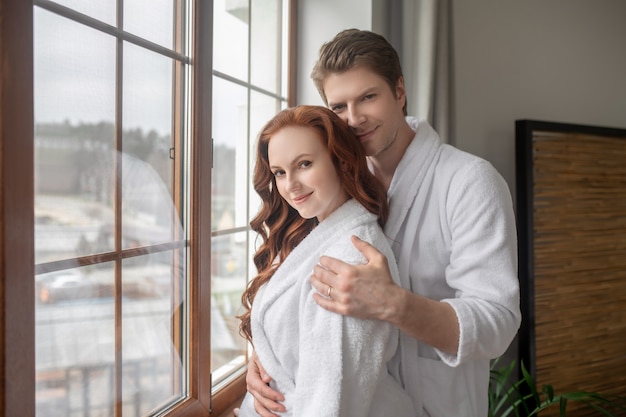 In love. A man and a woman in white bathrobes standing near the window and feeling in love