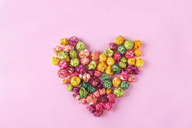 Love movies concept. Colorful rainbow caramel candy Popcorn scattered on pink background, heart shaped close up, copy space for text. Cinema snack concept