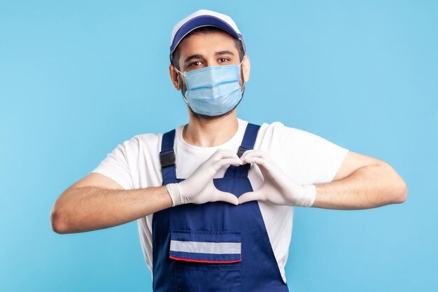 Love our customers. Handyman in overalls, mask and gloves showing heart gesture and smiling friendly to camera. Profession of service industry, courier delivery, housekeeping maintenance. isolated