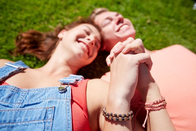 Photo love and people concept - close up of happy teenage couple lying on grass and holding hands with wristbands at summer