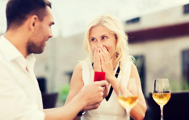 Foto concetto di amore, proposta, relazioni, persone e vacanze - coppia felice con anello di fidanzamento in una piccola confezione regalo rossa e bicchieri di vino sulla terrazza del ristorante