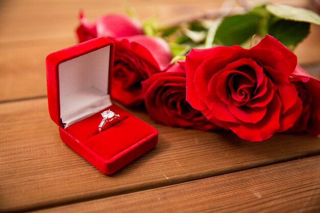 love, proposal, valentines day and holidays concept - close up of gift box with diamond engagement ring and red roses on wood